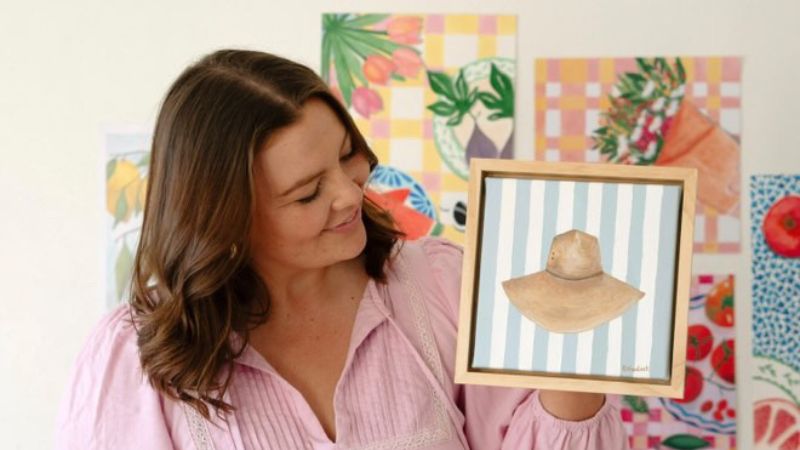 Woman holding framed artwork of a hat.